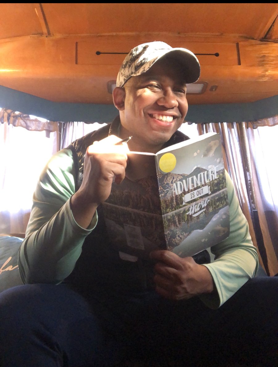 Greg McCoy smiling, holding an open notebook that reads Adventure Is Out There.